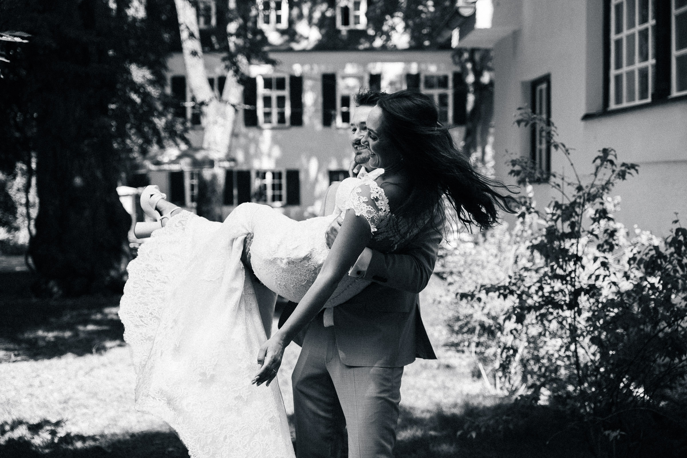 Schwarzweißaufnahme eines Hochzeitspaars: Der Bräutigam trägt die Braut im Freien